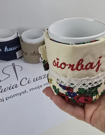 Ręka trzyma kubek z beżowym pokrowcem w kwiaty i koronką, z napisem "sionbaj". W tle inne realizacje.