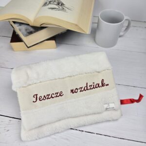 Biały puszysty otulacz na książkę z haftem "Jeszcze rozdział.." leży na stole obok otwartej książki i kubka.