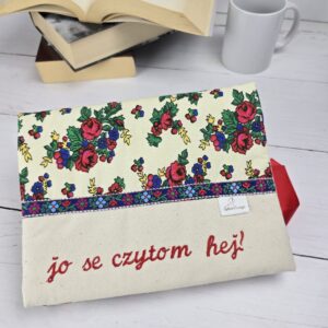 Otulacz na książkę z folkowym wzorem kwiatów i haftem "jo se czytom hej!" na jasnym tle, obok książki i kubek