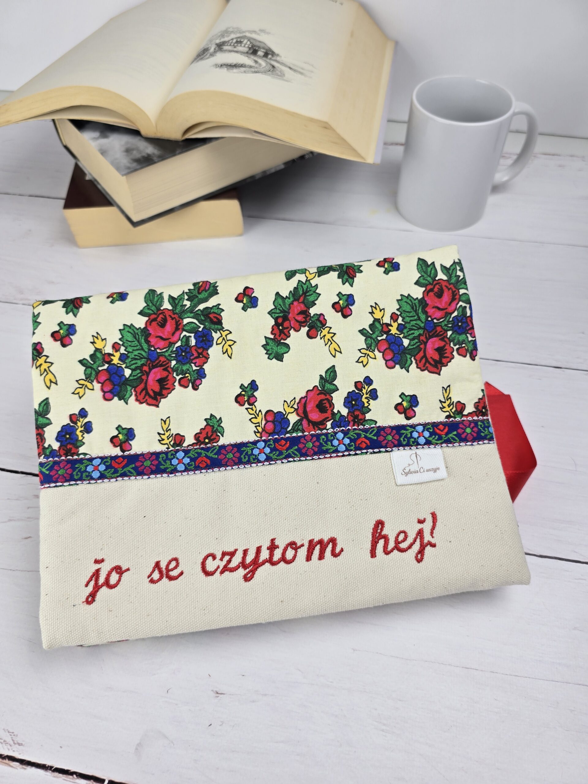 Otulacz na książkę z folkowym wzorem kwiatów i haftem "jo se czytom hej!" na jasnym tle, obok książki i kubek