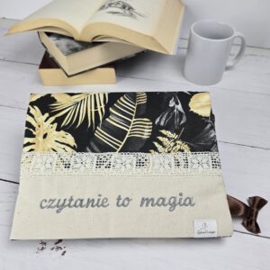 Otulacz na książkę z haftem "czytanie to magia", koronką i wzorem w liście, obok stos książek i kubek
