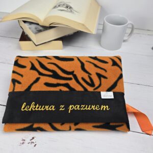 Otulacz na książkę w tygrysie paski z haftem "lektura z pazurem", obok otwarte książki i kubek