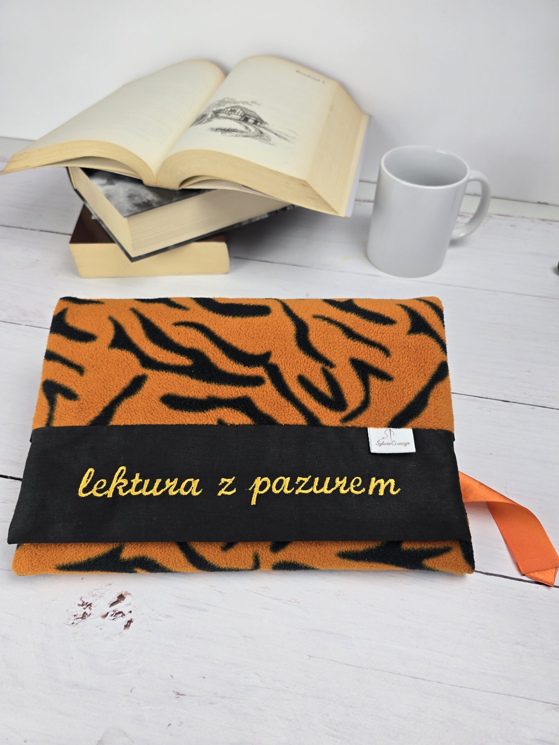 Otulacz na książkę w tygrysie paski z haftem "lektura z pazurem", obok otwarte książki i kubek