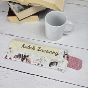 Otulacz na kubek z haftem "kubek Zuzanny", biały ceramiczny kubek i otwarte książki na drewnianym blacie.