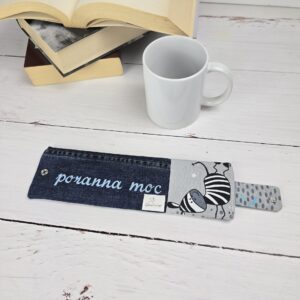 Otulacz na kubek z haftem "poranna moc" i wzorem w pszczoły, obok biały kubek i książki