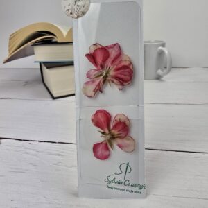 Biała zakładka do książki z dwoma suszonymi kwiatami pelargonii, leżąca na białym drewnianym stole obok książek.